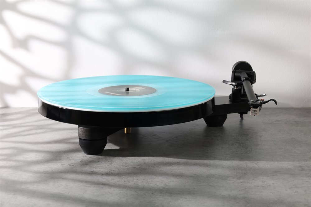 The Rega Replay in matt black at home on a stone surface, a turquoise record on the platter, leaf shadows on the wall behind.