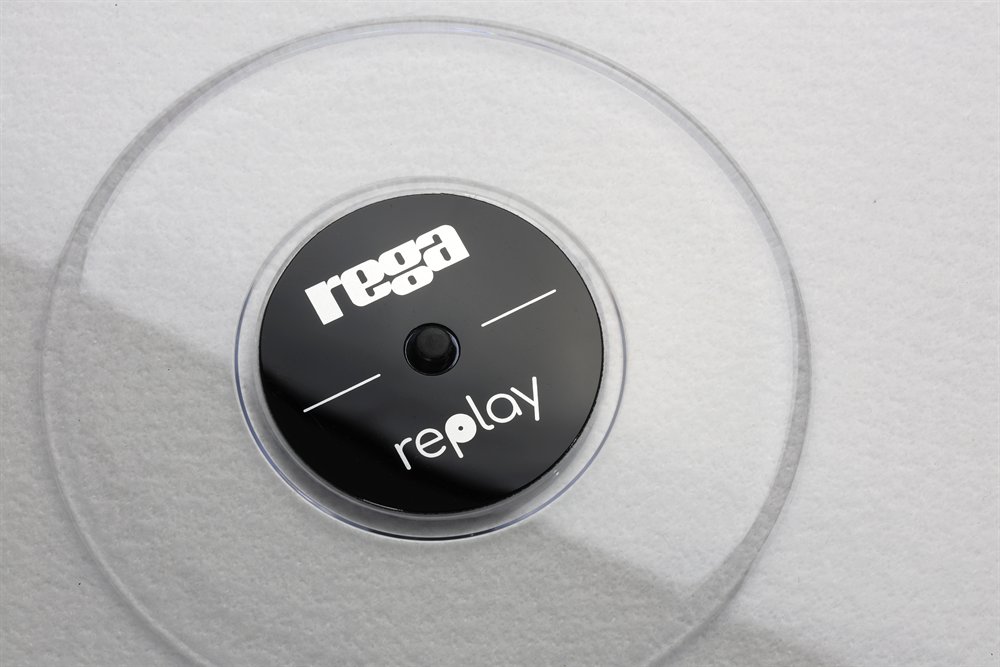 Detail of the Rega Replay badge: the black centre disc with the Rega and Replay logos, set in clear acrylic over a white mat.